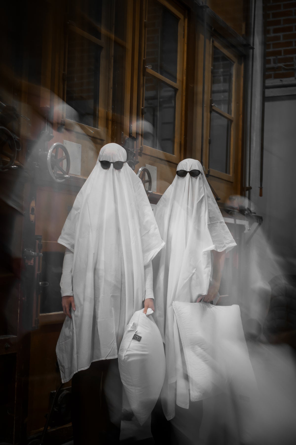 Two people wearing white ghost costumes with black sunglasses stand in front of a building with dark windows, surrounded by fog and motion-blurred overlays.