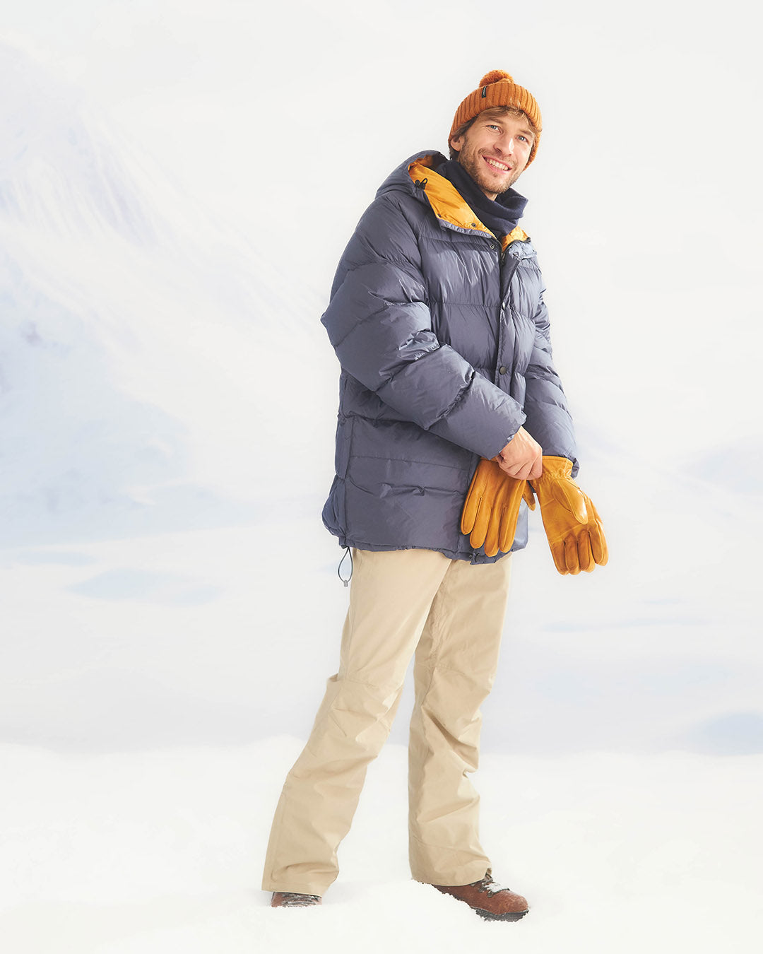 Person wearing a blue down coat , beige pants, and yellow gloves against a white background