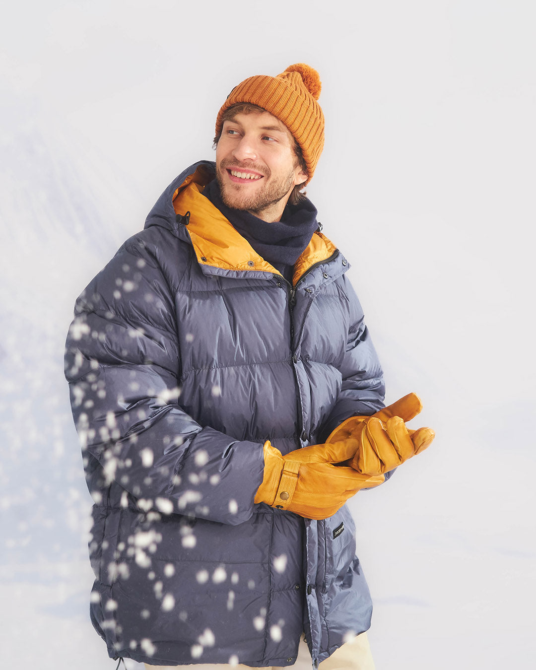 Man wearing a blue down coat with yellow lining, orange beanie, and gloves standing in snow.