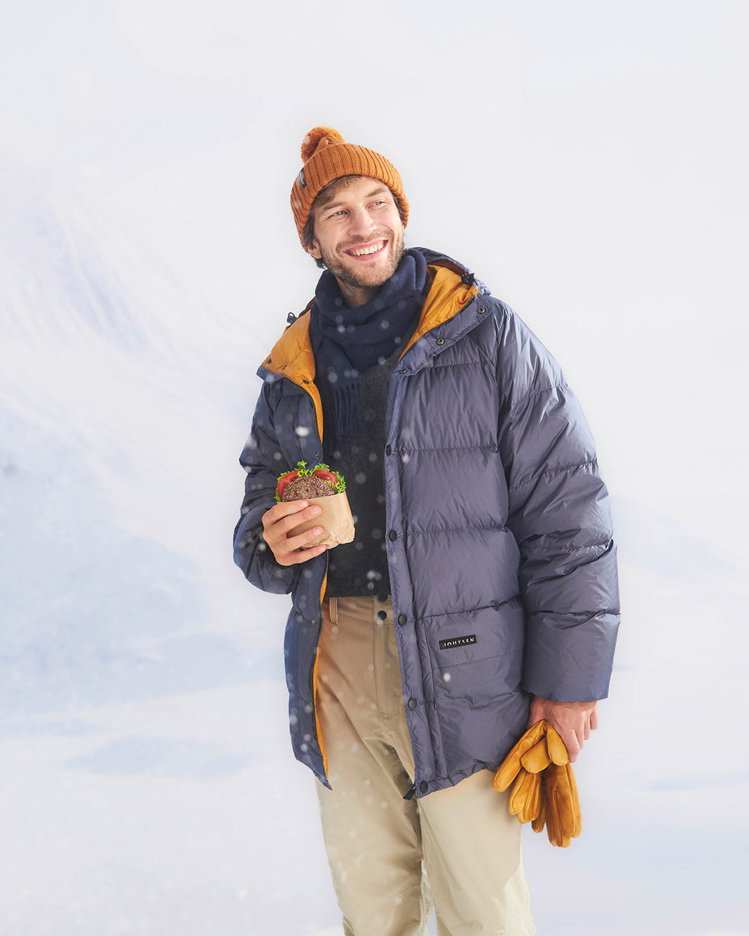 Man wearing a blue down coat and orange beanie holding a sandwich against a light blue background