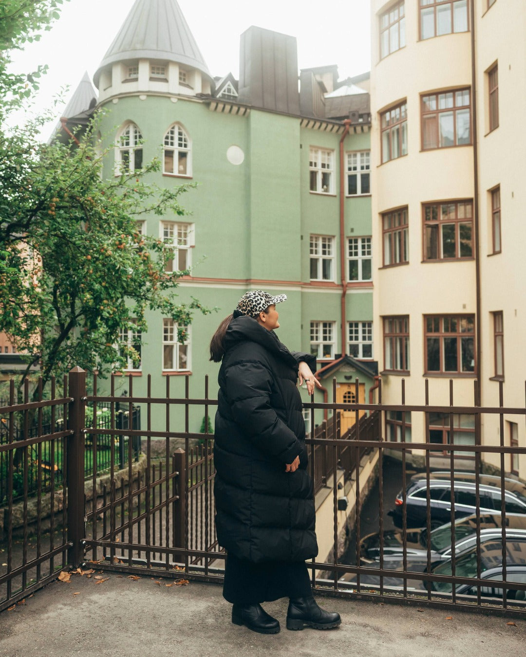 Person standing on a balcony wearing a long down coat with colorful buildings in the background