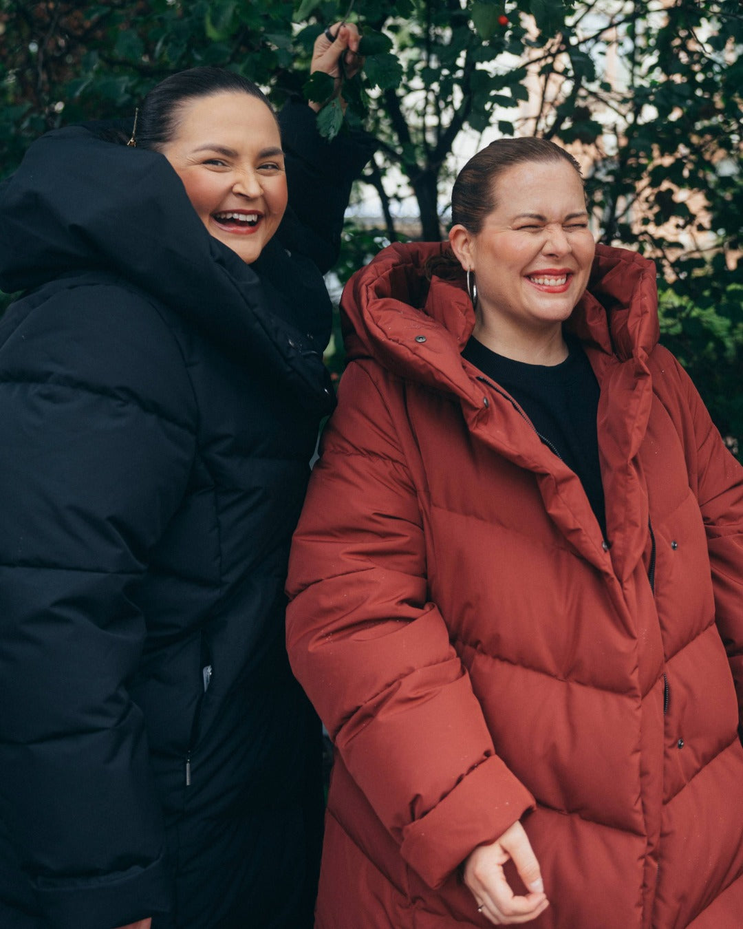 Two people outdoors, one in a black coat and the other in a red coat, with trees in the background.
