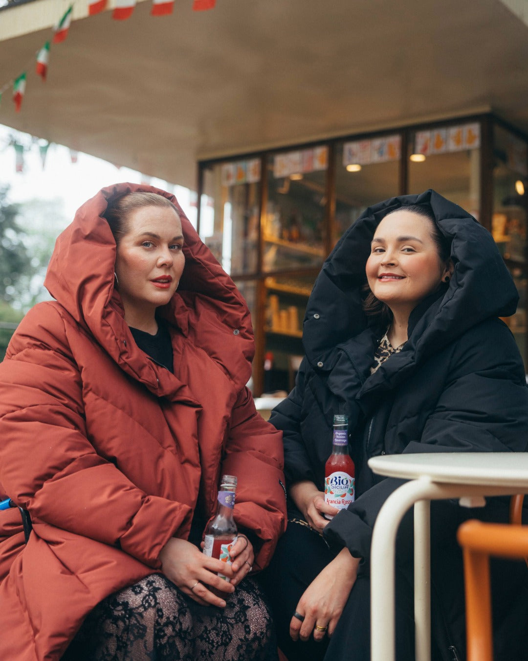 Two women sitting outdoors in winter coats with drinks.