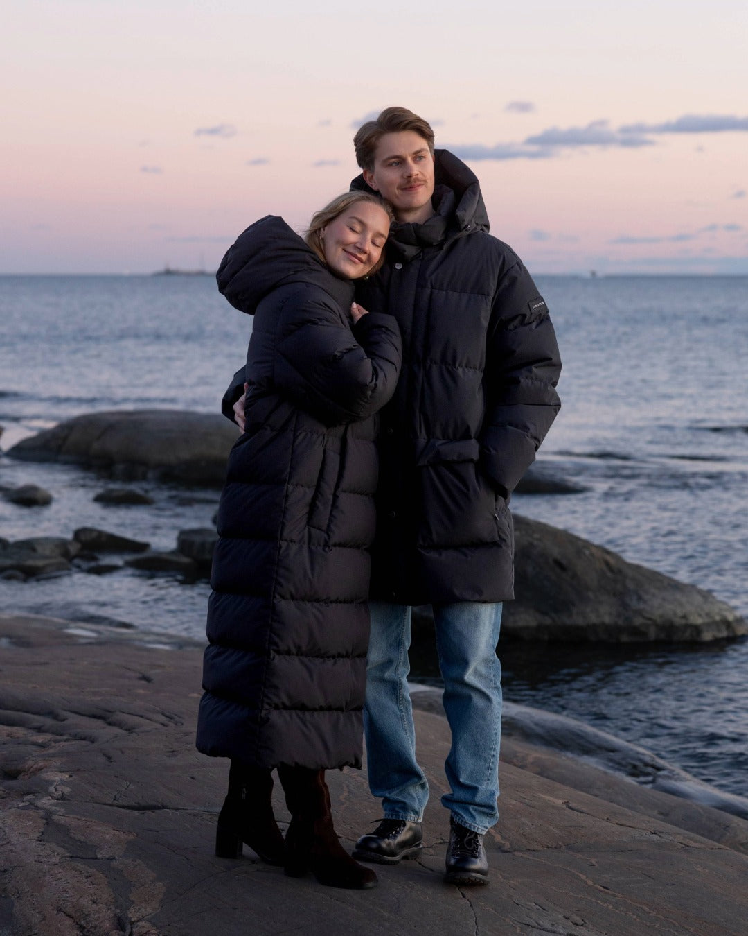 Two people in long black down coats standing on a beach at sunset.