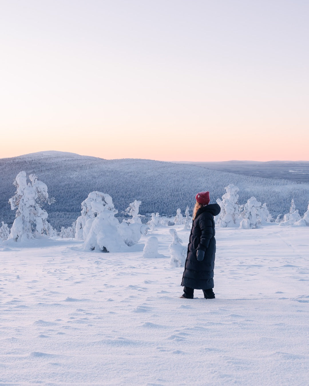 Person standing in a snowy landscape with snow-covered trees and mountains in the background. She is wearing a black (color: Black bird) Blenda down coat.