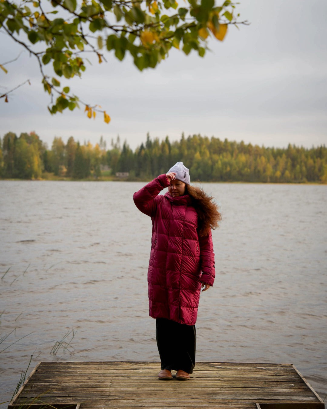 Person in a dark red coat standing on a wooden dock by a lake with trees in the background