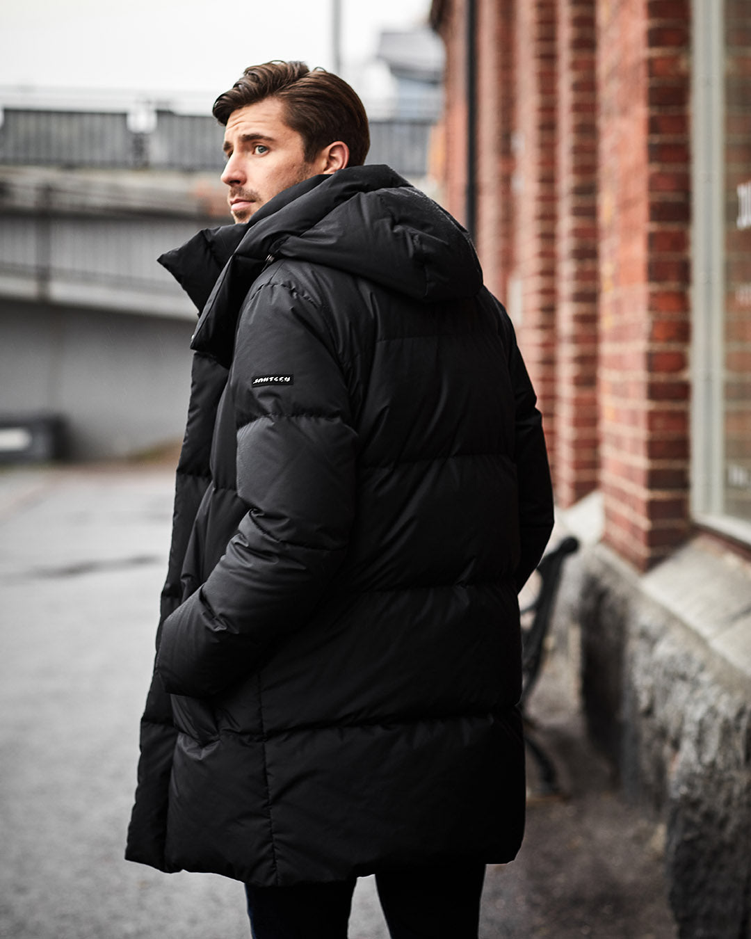 Man wearing a black down coat standing on a street with a brick building in the background
