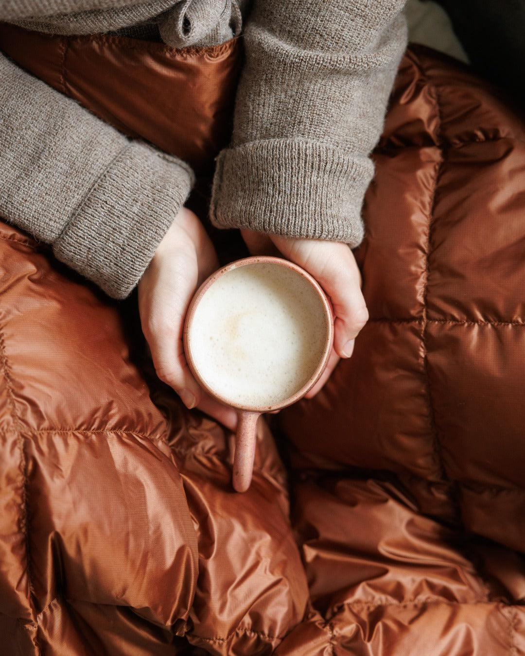 Person holding a mug of coffee or tea in a cozy setting with brown (color: chocolate mousse) Kulkuri down blanket