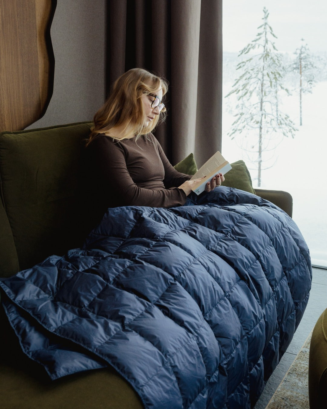 Woman reading a book under a blue (color: Honeyberry) down blanket in a cozy room with large windows.