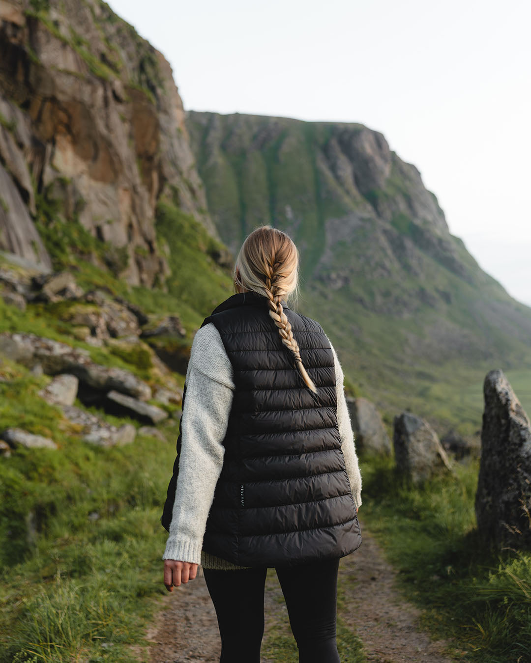 A woman stands with her back on a mountain trail. She is wearing a black Lady down vest (color: charcoal black), a grey knit sweater, and black trousers. Her hair is tied loosely in a braid.