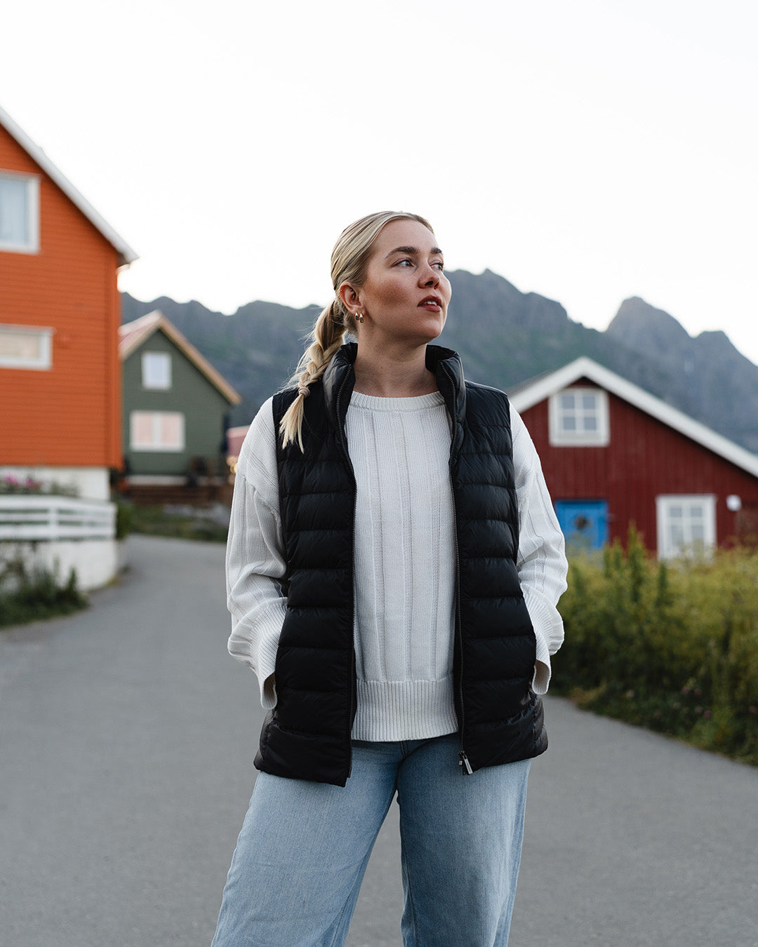 A woman stands in a fishing village, looking to the left. She is wearing a black horizontally quilted Lady down vest (color: charcoal black), a white knit sweater, and light blue jeans. The vest is unzipped.