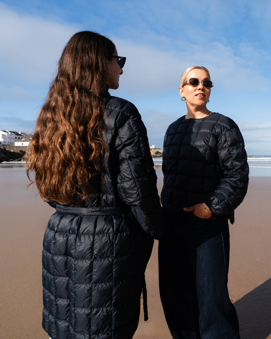 Two women in black down coats standing on a beach with a clear blue sky.