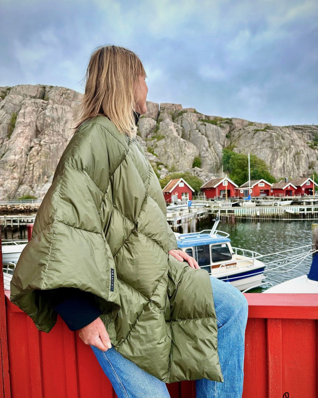 Person wearing a green Otava down poncho sitting by a harbor with red houses and rocky cliffs in the background.