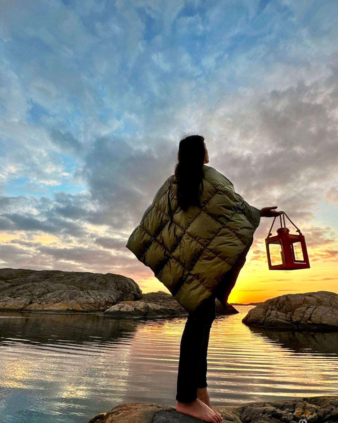 Person in a green Otava down poncho holding a lantern by a body of water at sunset.
