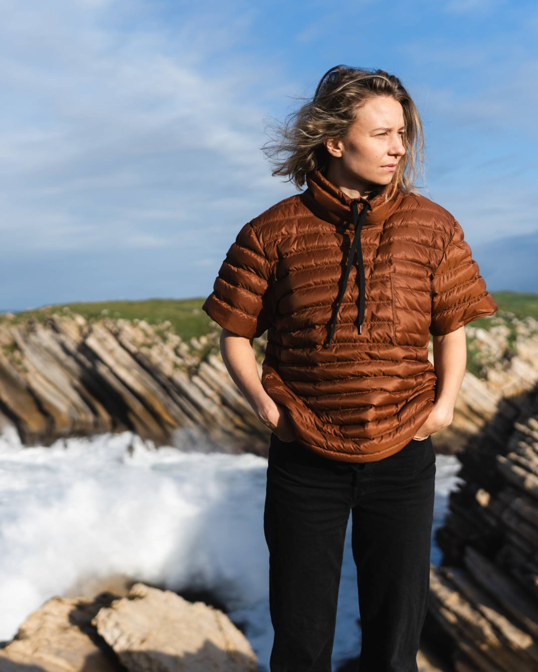 Person wearing a brown down shirt standing on a coastal landscape with ice and rocks.