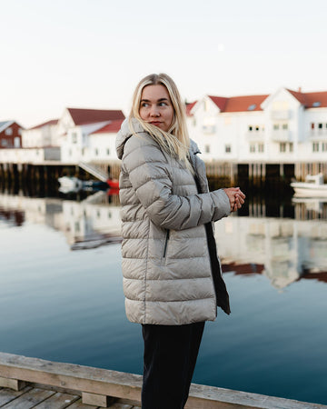 A woman stands on a pier by the water, with light-colored houses and boats of a fishing village in the background. She is wearing a beige Ruut light down coat (color: Champagne) with a hood and zippered pockets.