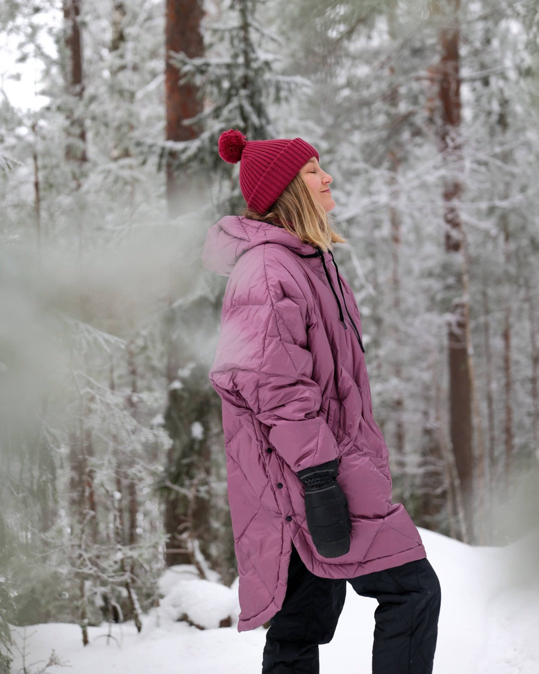 Person wearing a light pink down anorak and red beanie standing in a snowy forest