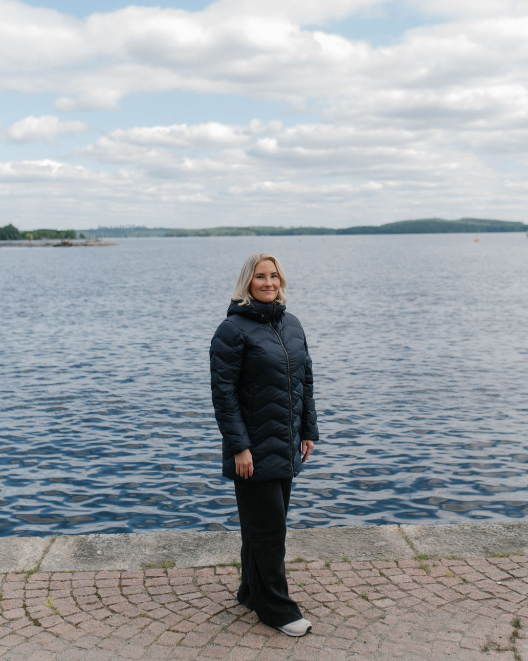 A woman wearing a navy blue (color: Night sky) quilted light down coat and black pants stands on a stone walkway next to a large body of water under a cloudy sky.