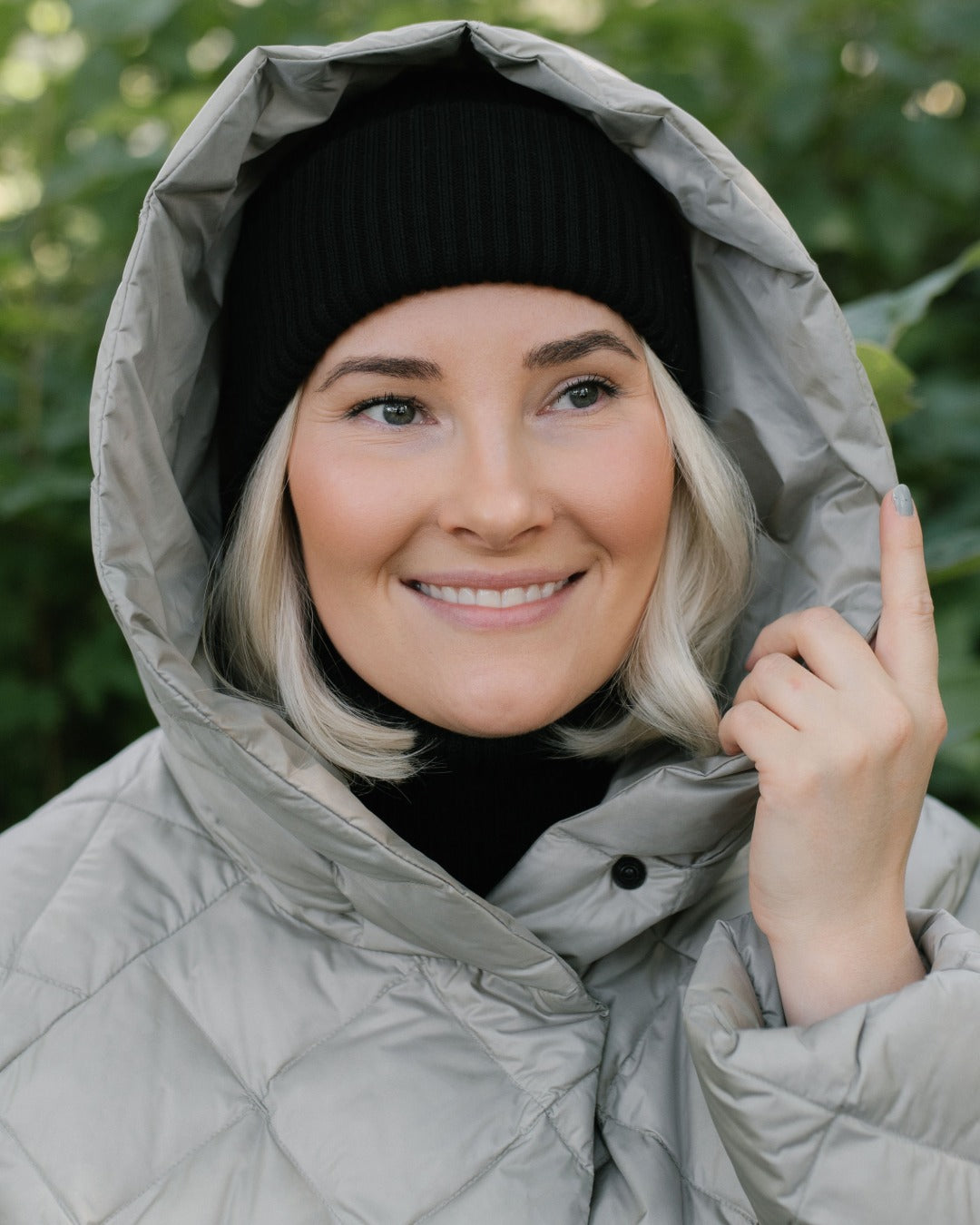 A smiling woman wearing a light gray (color: Champagne) hooded light down coat and a black knit beanie, photographed outdoors with blurred green foliage in the background.