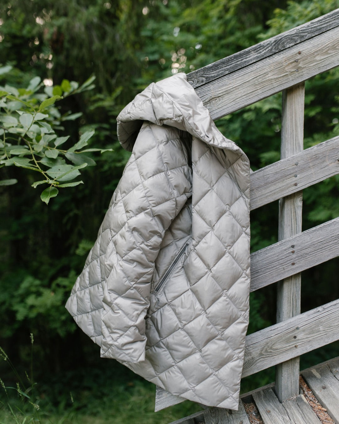 A light gray (color: Champagne) diamond-quilted light down coat with a hood, draped over a weathered wooden fence in an outdoor garden setting.