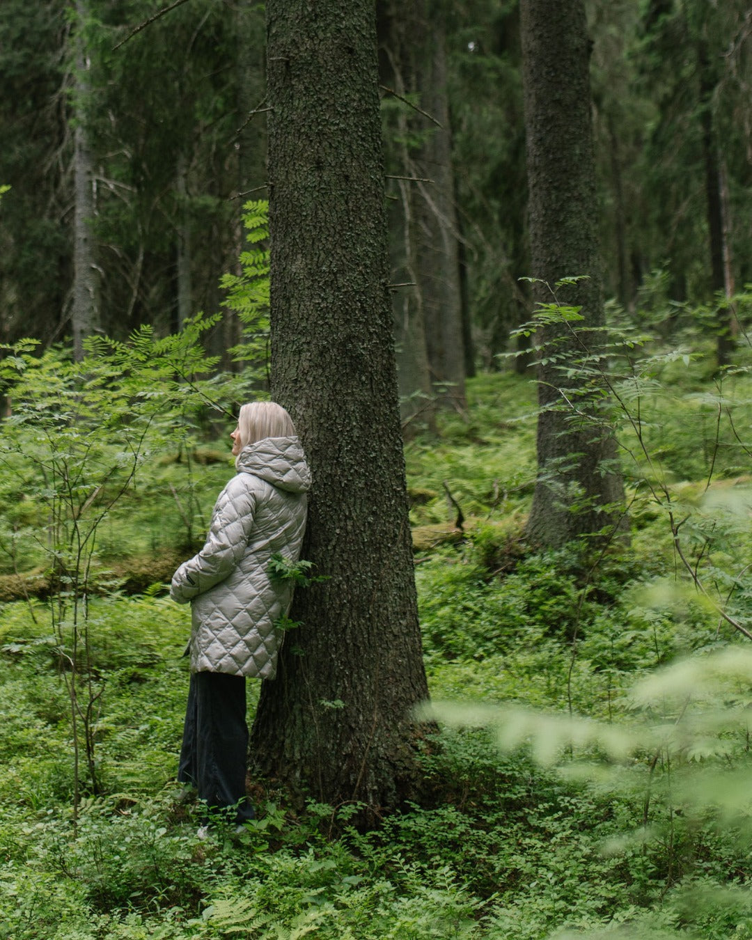 A woman in a light gray (color: Champagne) quilted light down coat and dark jeans stands against a large tree trunk in a lush green forest.