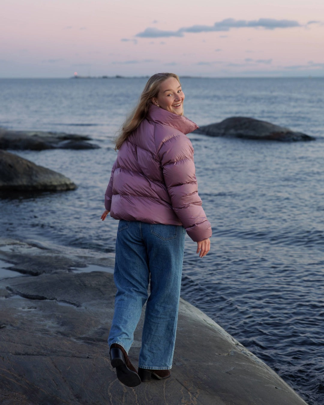 Person wearing a pink down coat standing on a rocky shoreline with water and sky in the background