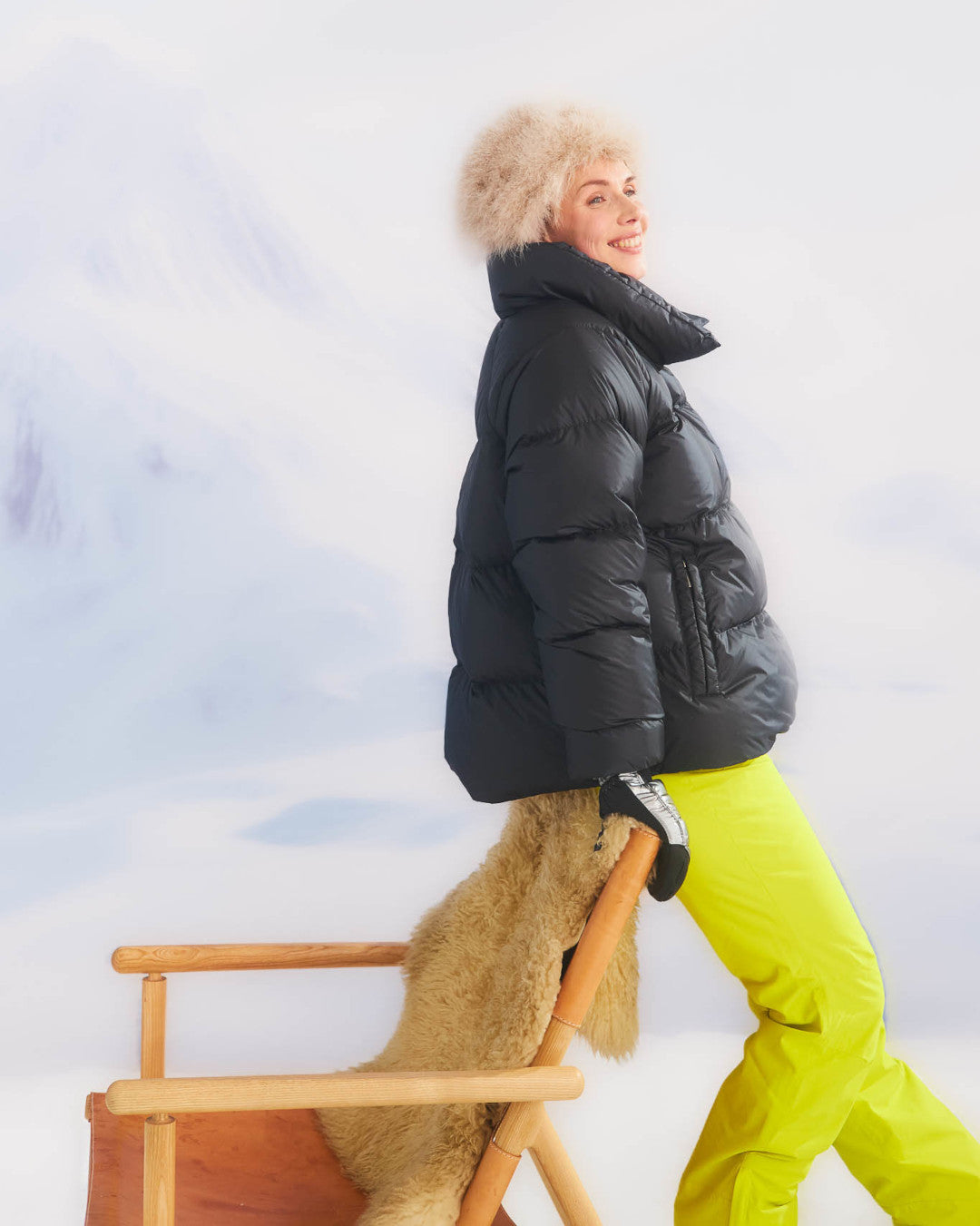 Person wearing a black down coat and yellow pants sitting on a wooden chair with a fur throw against a snowy background.