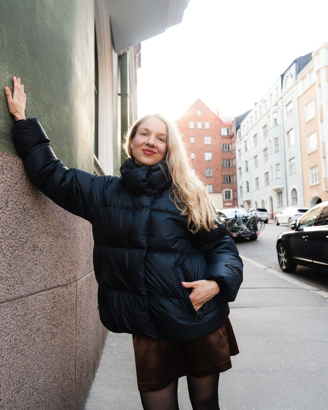 A woman with blonde hair wearing a black down coat, brown skirt, and black tights stands on a city street, leaning her hand against a dark green wall.