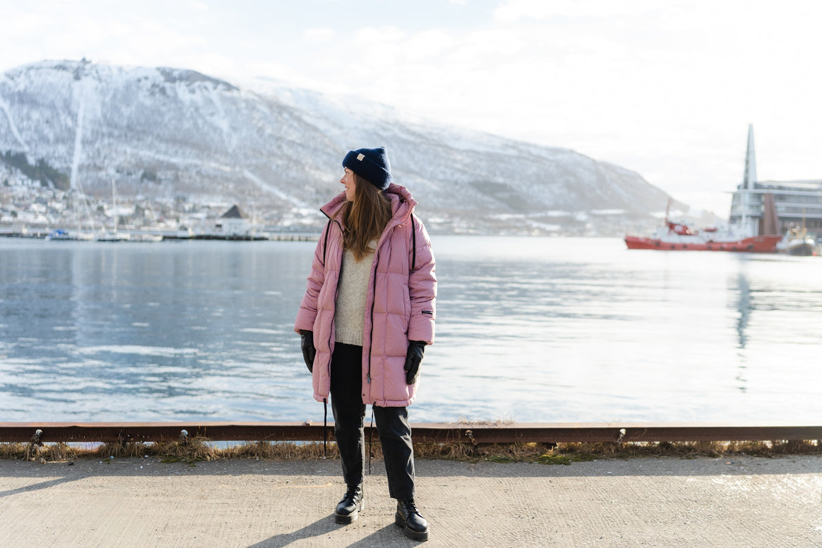 A woman stands on a waterfront promenade wearing a pink down coat, black beanie, black pants, and black gloves, with a snowy mountain and harbor in the background.