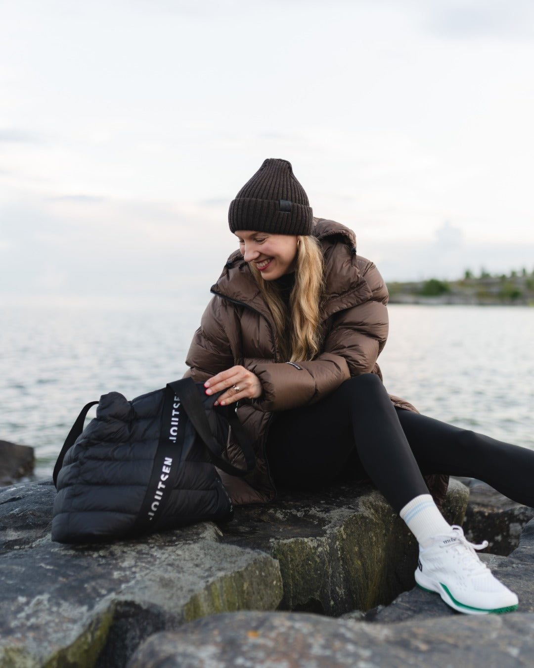 A woman sitting on rocks by the water wearing a brown down coat, black beanie, black leggings, and white sneakers, next to a black puffer totebag