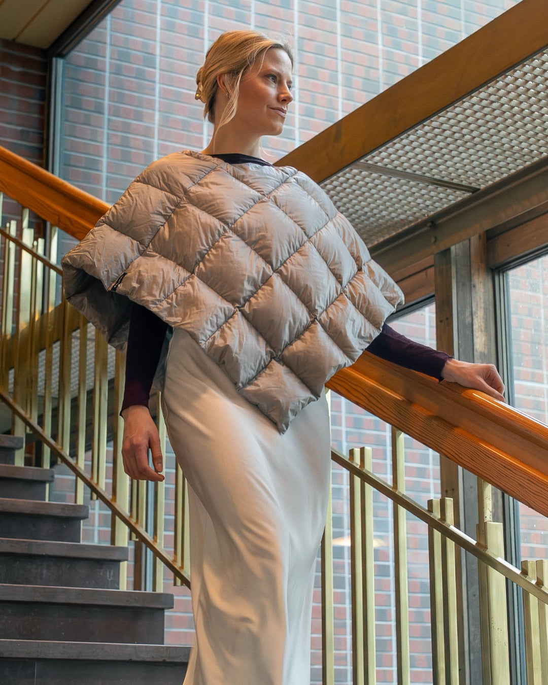 Woman wearing a quilted light gray Unelma down poncho in an indoor setting with wooden railings and glass windows.
