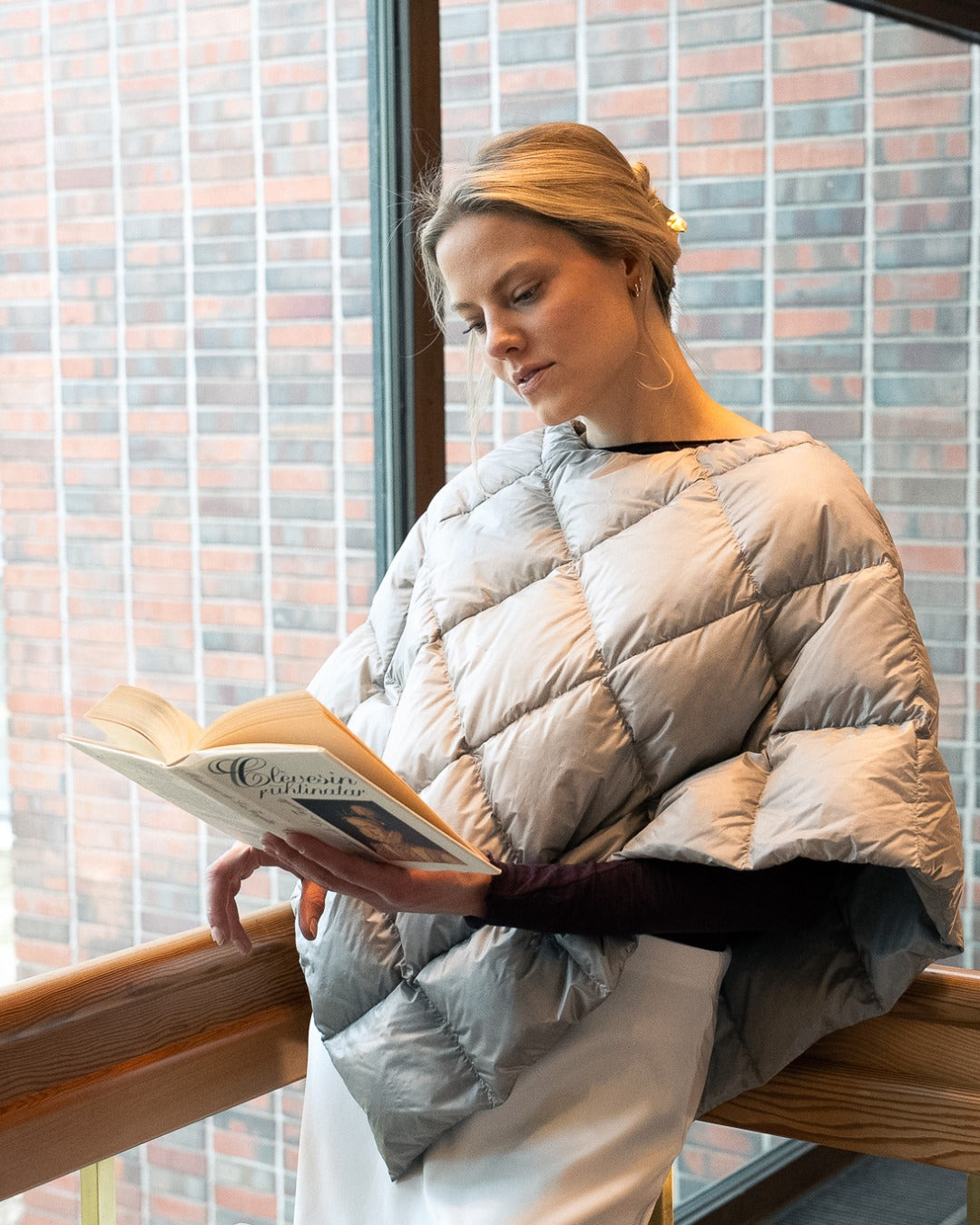 Woman reading a book wrapped in a light Unelma down poncho by a window.