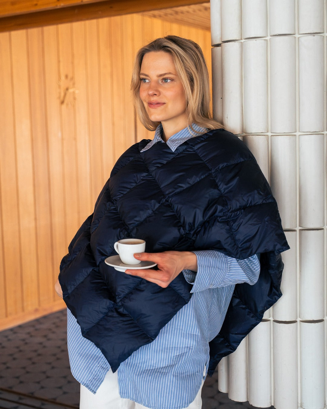 Woman wearingdark blue Unelma down poncho and holding a cup of coffee against a tiled wall.