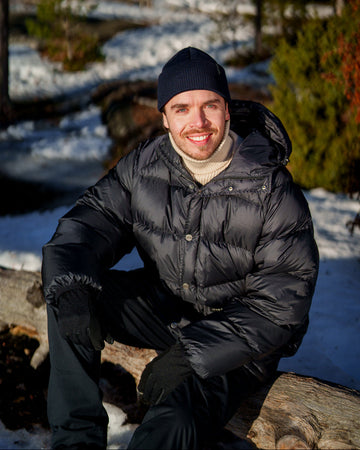 Man wearing a charcoal black Tauko down coat with horizontal quilting and a hood. The coat has a front button closure and is styled with a knit beanie, gloves, and dark trousers.