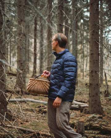 A man is standing in the forest holding a woven basket in his hand. He is wearing a blue Hile light down coat (color: Storm).