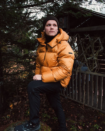 Person wearing an orange down coat standing outdoors with trees and a building in the background