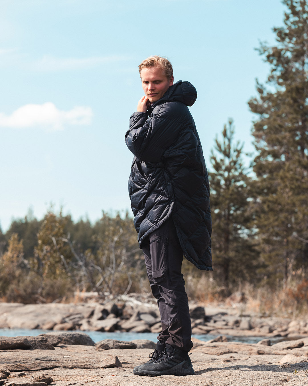 Person wearing a black Velho down anorak and pants standing on a rocky shoreline with trees in the background.