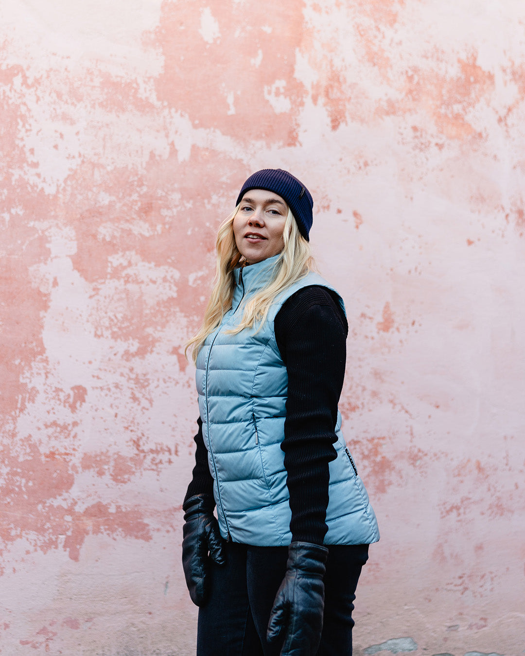 A woman stands in front of a pink wall wearing a blue (color: Blue Bell) Lady down vest. The vest has a lightly shiny surface and horizontal quilting. Her outfit also includes a black beanie, sweater, trousers, and leather gloves.