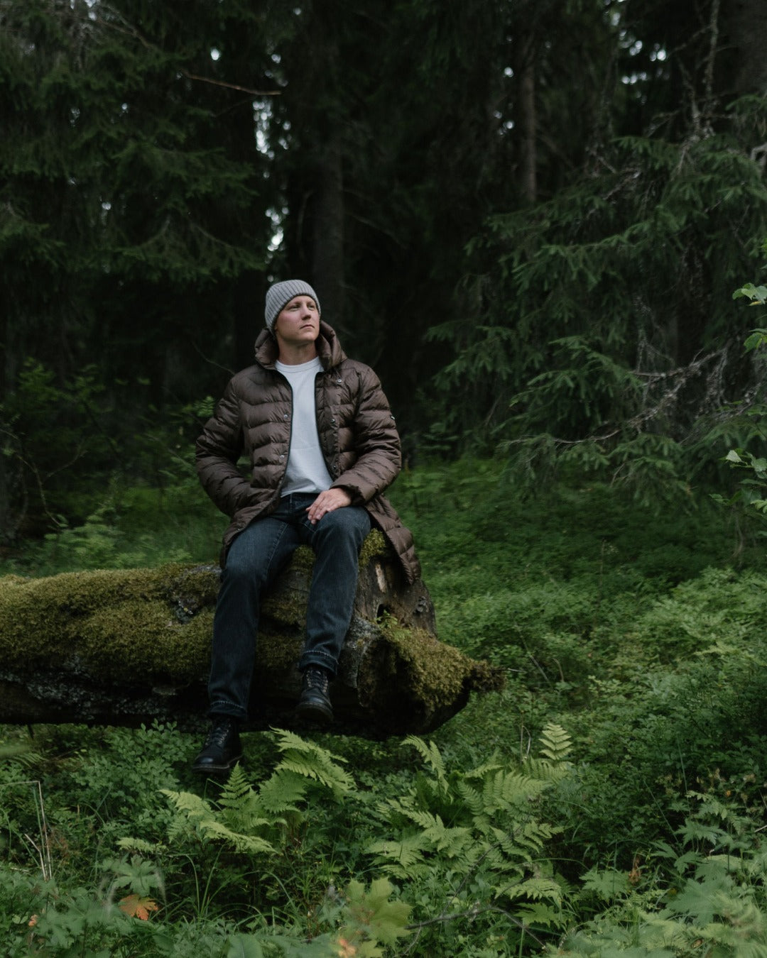 A man wearing a brown down coat, white t-shirt, dark jeans, and a gray beanie sits on a mossy log in a dense green forest.
