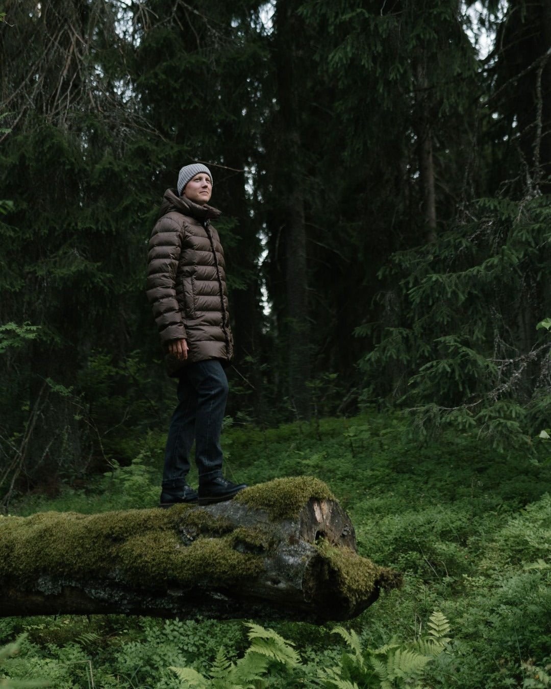 A man wearing a brown (color: Coffee quarts) light down coat, dark pants, and a gray beanie stands on a moss-covered log in a dense green forest.