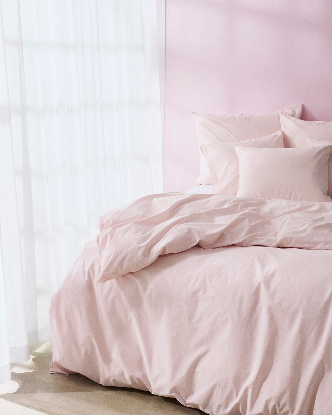 Pink bedding set on a bed with a light pink wall and white curtains.