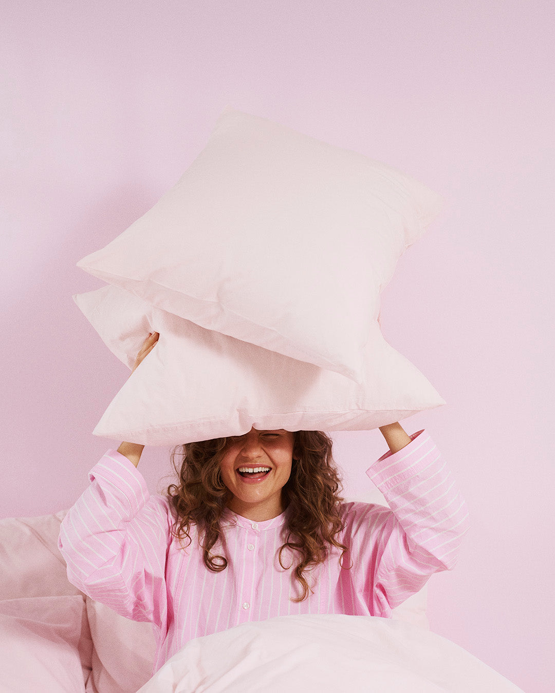 Person in pink pajamas holding white pillows against a light pink background