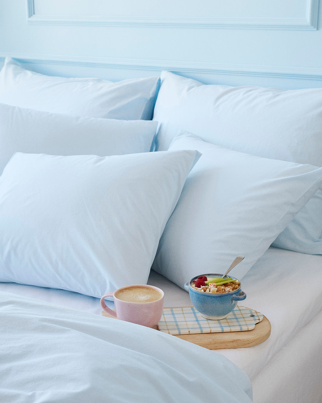 Light blue pillows on a bed with a small table holding a cup and bowl of fruit.