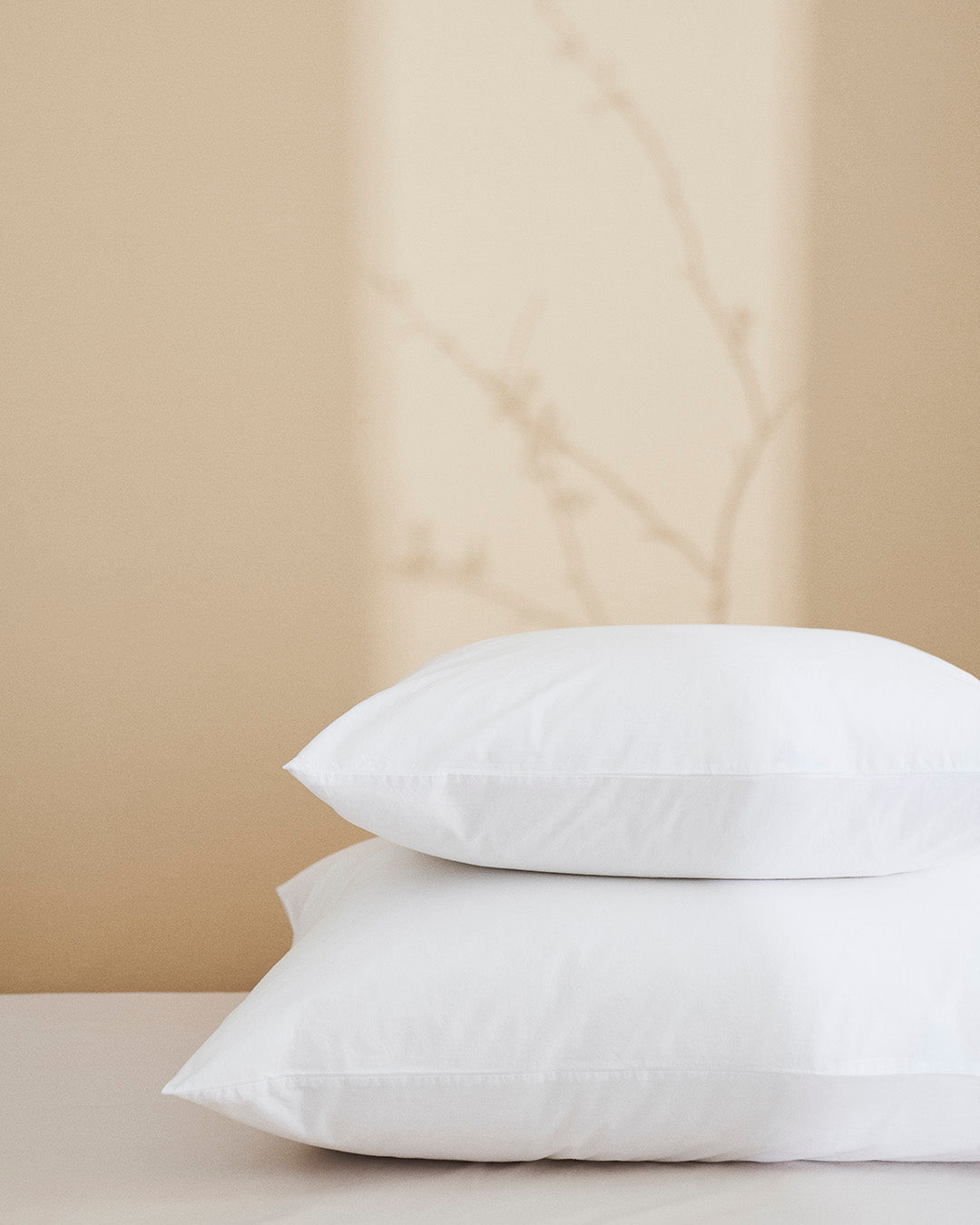 Two white pillows stacked on a beige surface with a beige curtain in the background