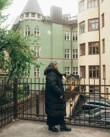 Person standing on a balcony wearing a long down coat with colorful buildings in the background