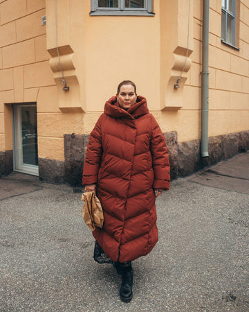 Person wearing a long red down coat standing on a street with a yellow building in the background.