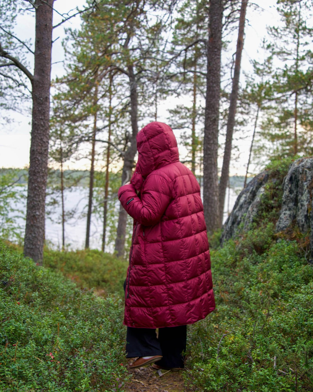 Person wearing a dark red light down coat standing in a forest by a lake