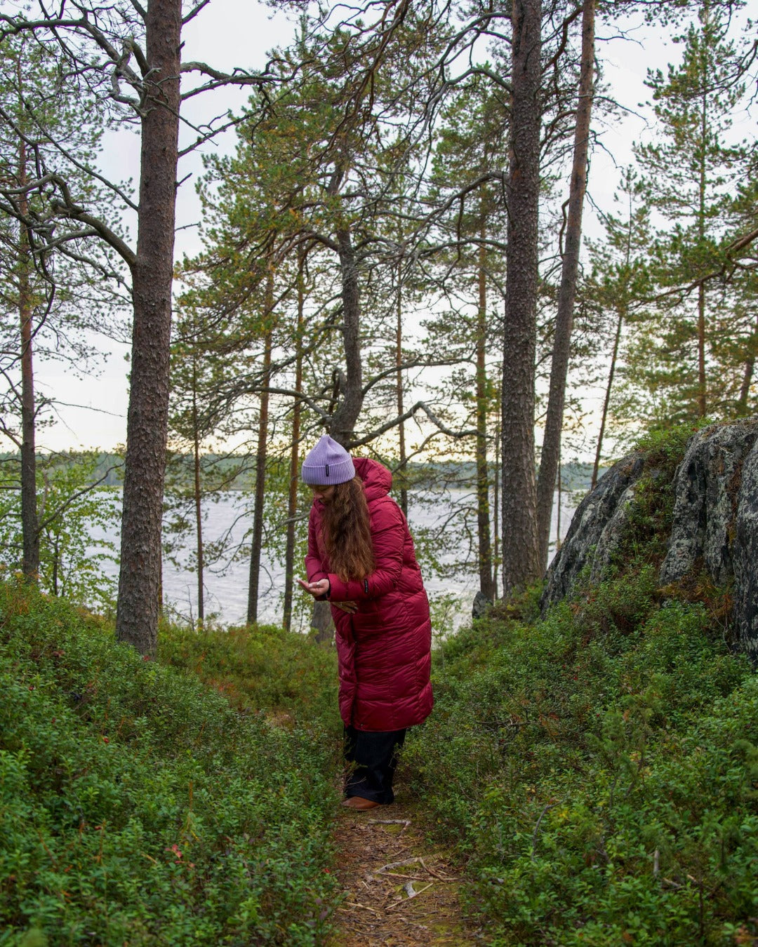 Person in a red coat standing on a path in a forest with a lake in the background