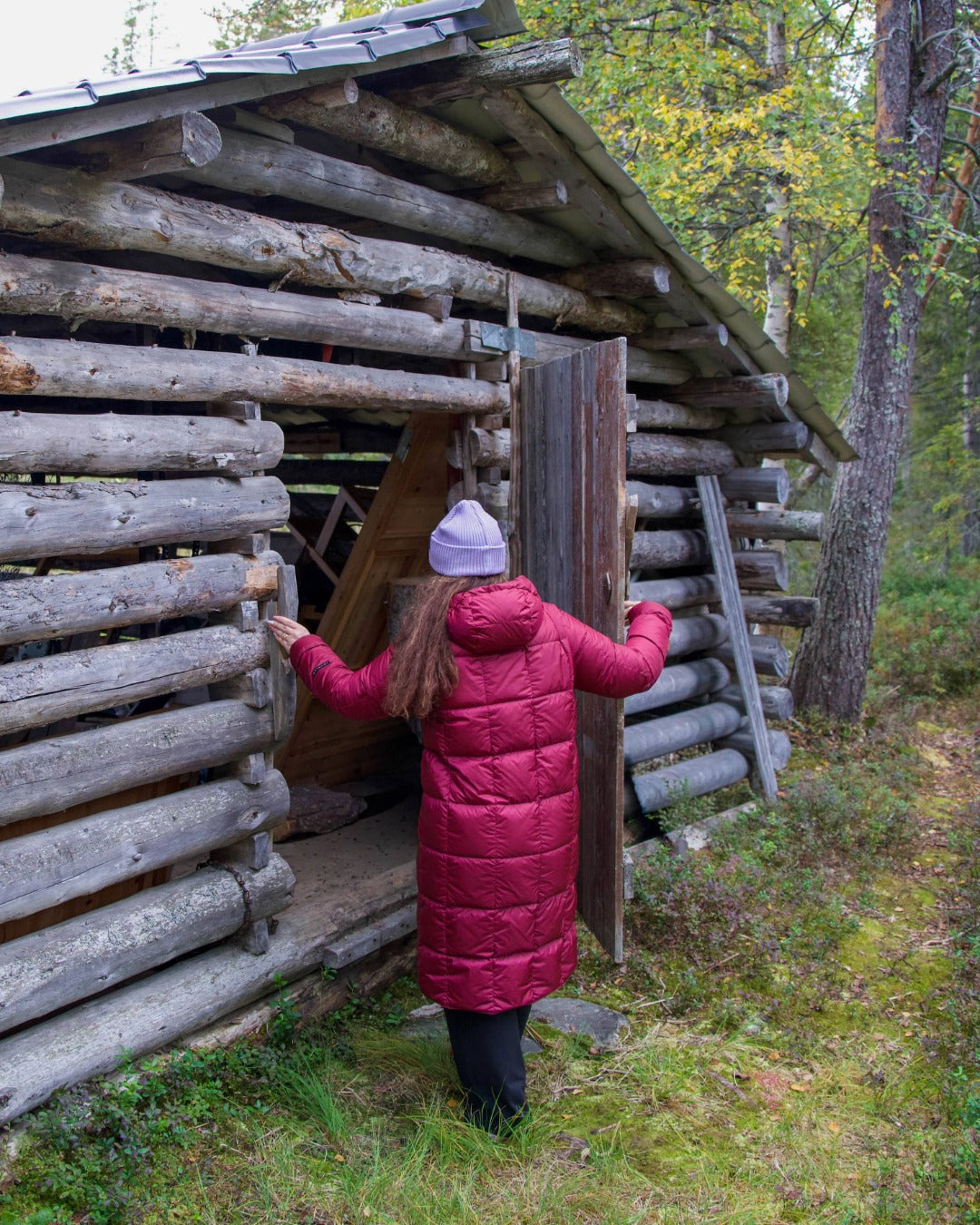 Person in a dark red coat standing in front of a wooden cabin in a forest setting