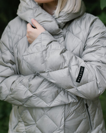A close-up of a person wearing a light gray (color: Champagne) quilted light down coat with a high collar and a small black brand patch on the sleeve that reads JOUTSEN.